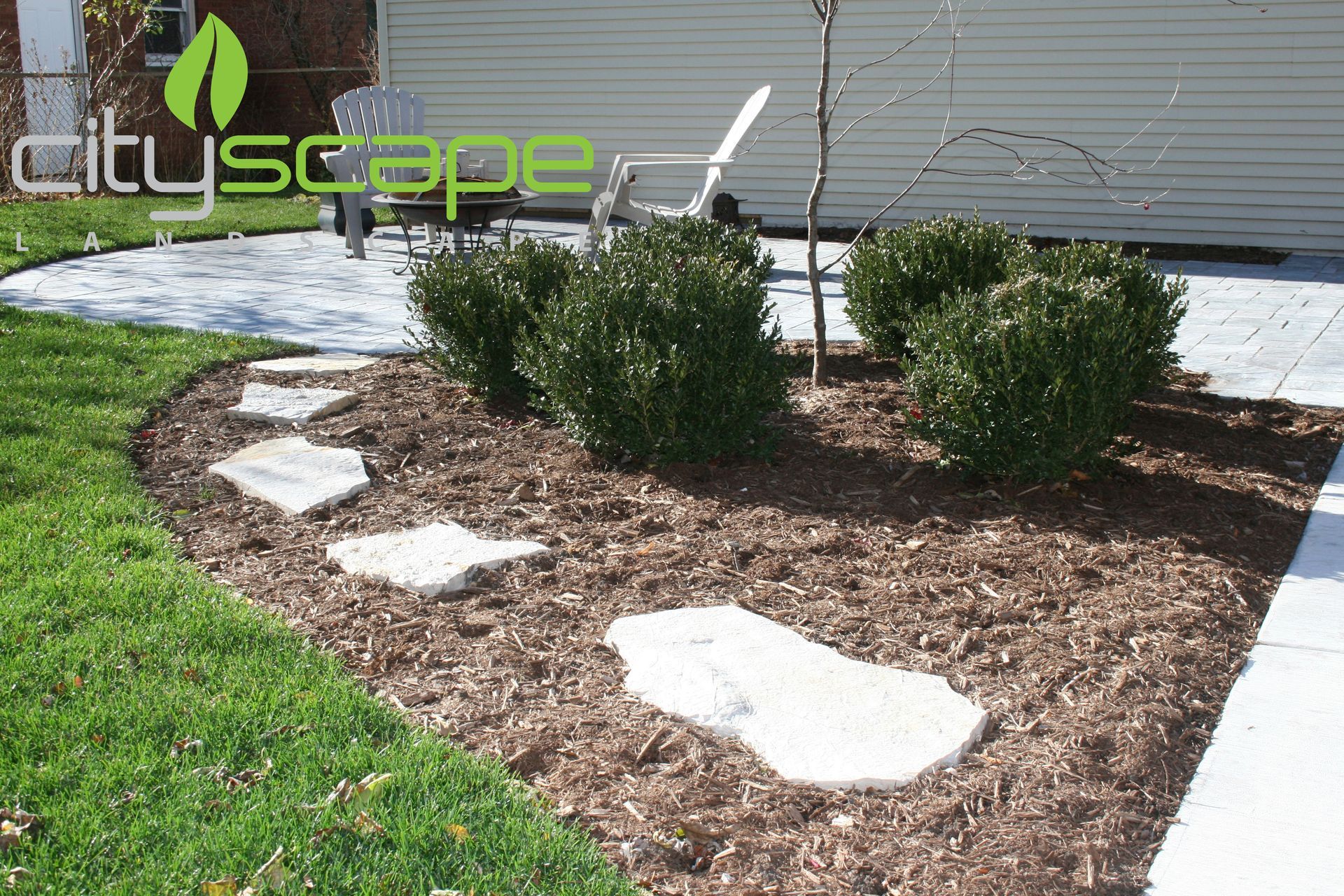 A landscaped garden bed with stepping stones, mulch, and green bushes, adjacent to a grassy lawn.