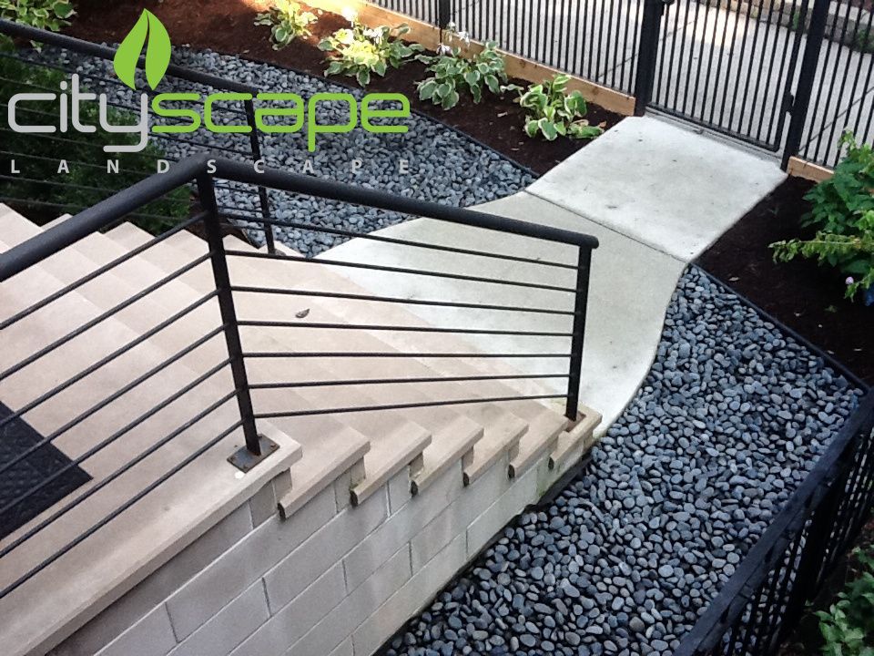 Modern outdoor staircase with black railing, concrete path, and dark stone ground cover.