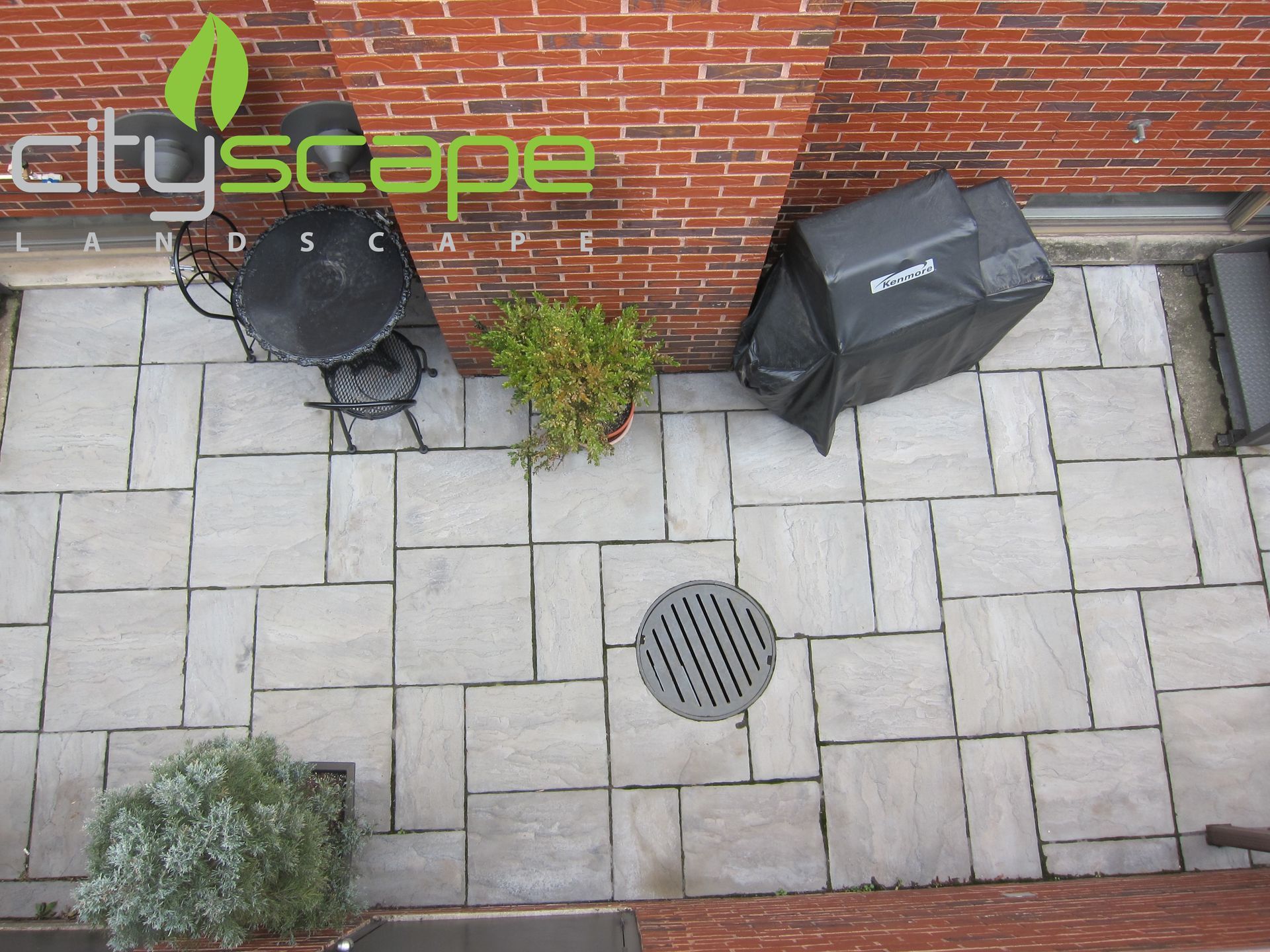 Overhead view of patio with brick wall, black grill cover, table, plants, and square paving stones.