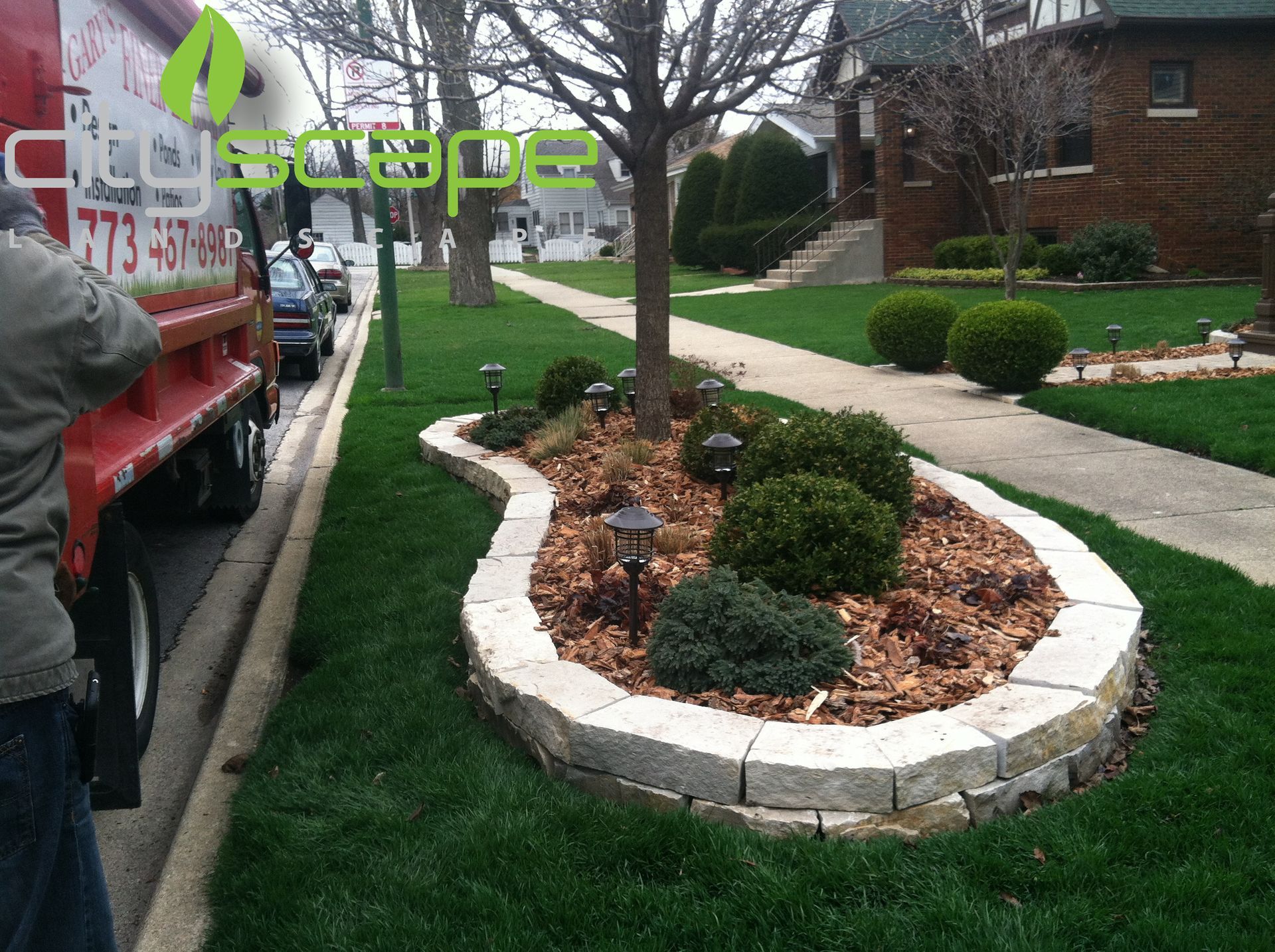 Landscaped garden bed with stone border, small shrubs, and tree on a lawn beside a street.
