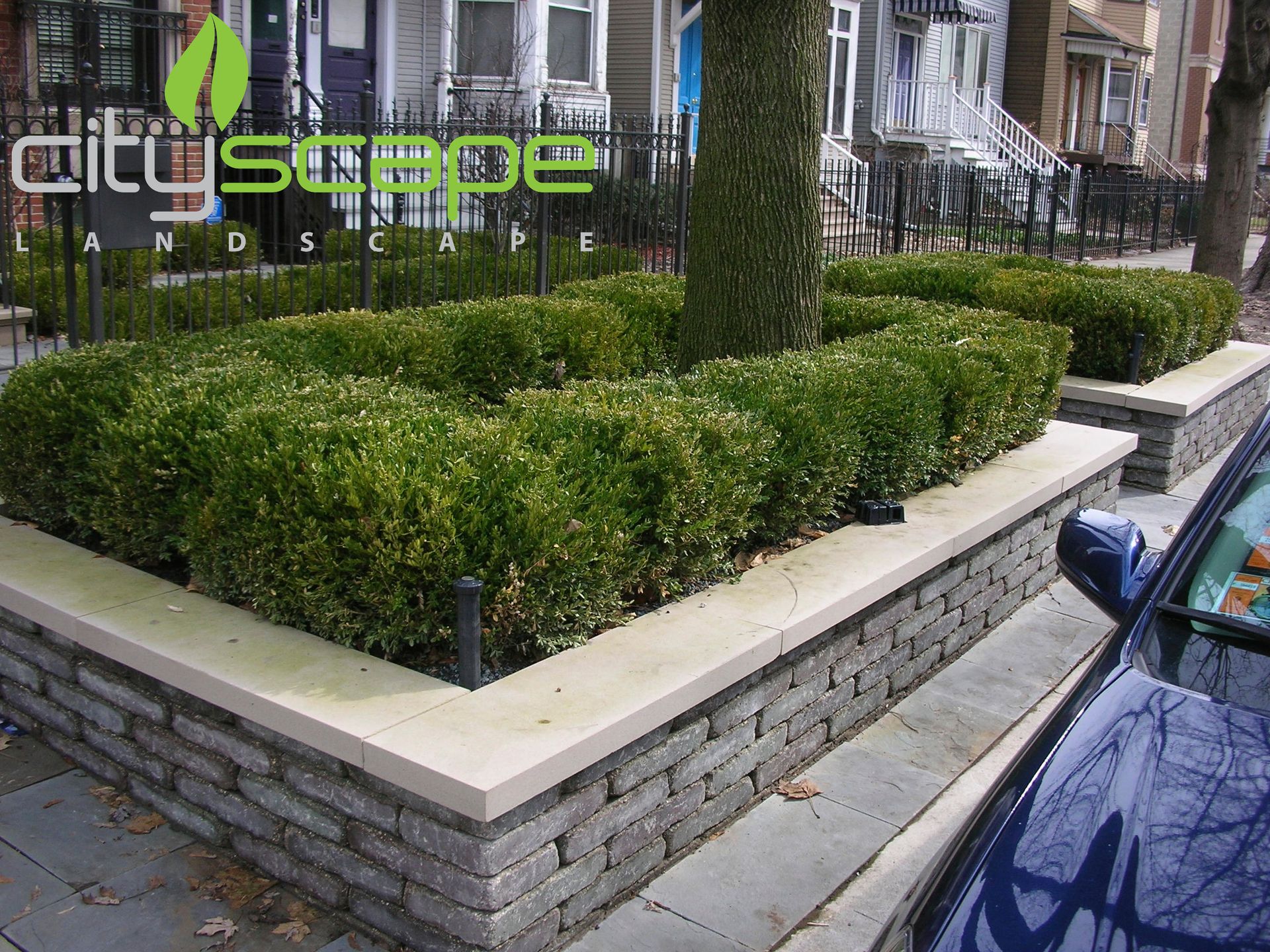 Green shrubs in a stone-walled planter on a city sidewalk. The landscaping company logo is in the top left.