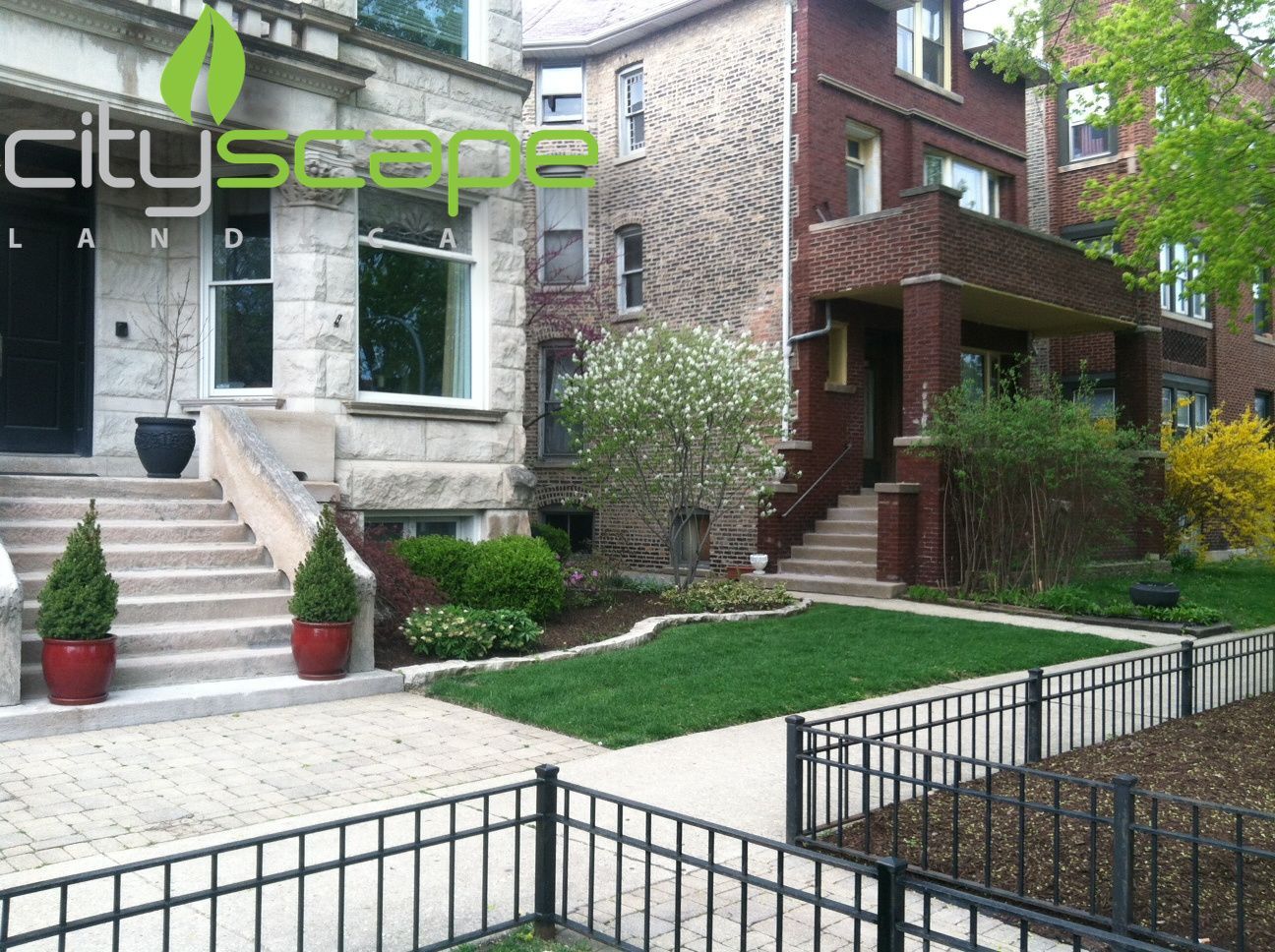 Two row houses with manicured lawns, landscaping, and a wrought-iron fence.