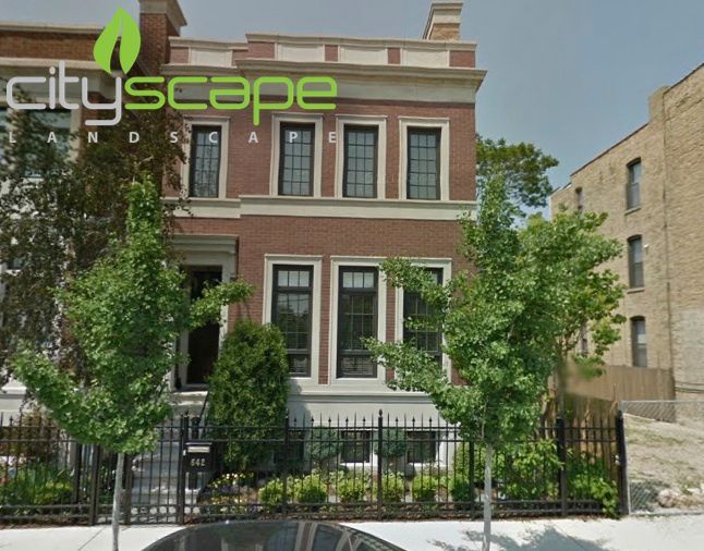 Two-story brick building with black windows, a black fence, and small trees in front.