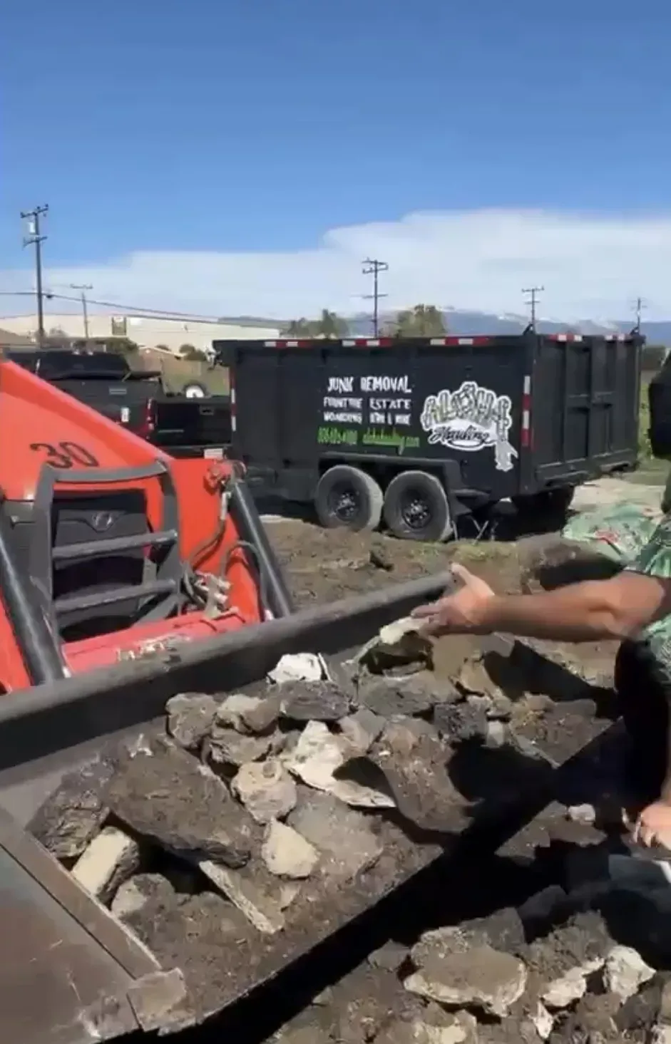 An orange skid steer scoop filled with concrete rubble near a black dump trailer in an outdoor lot of Happy Place liquor under a clear sky.