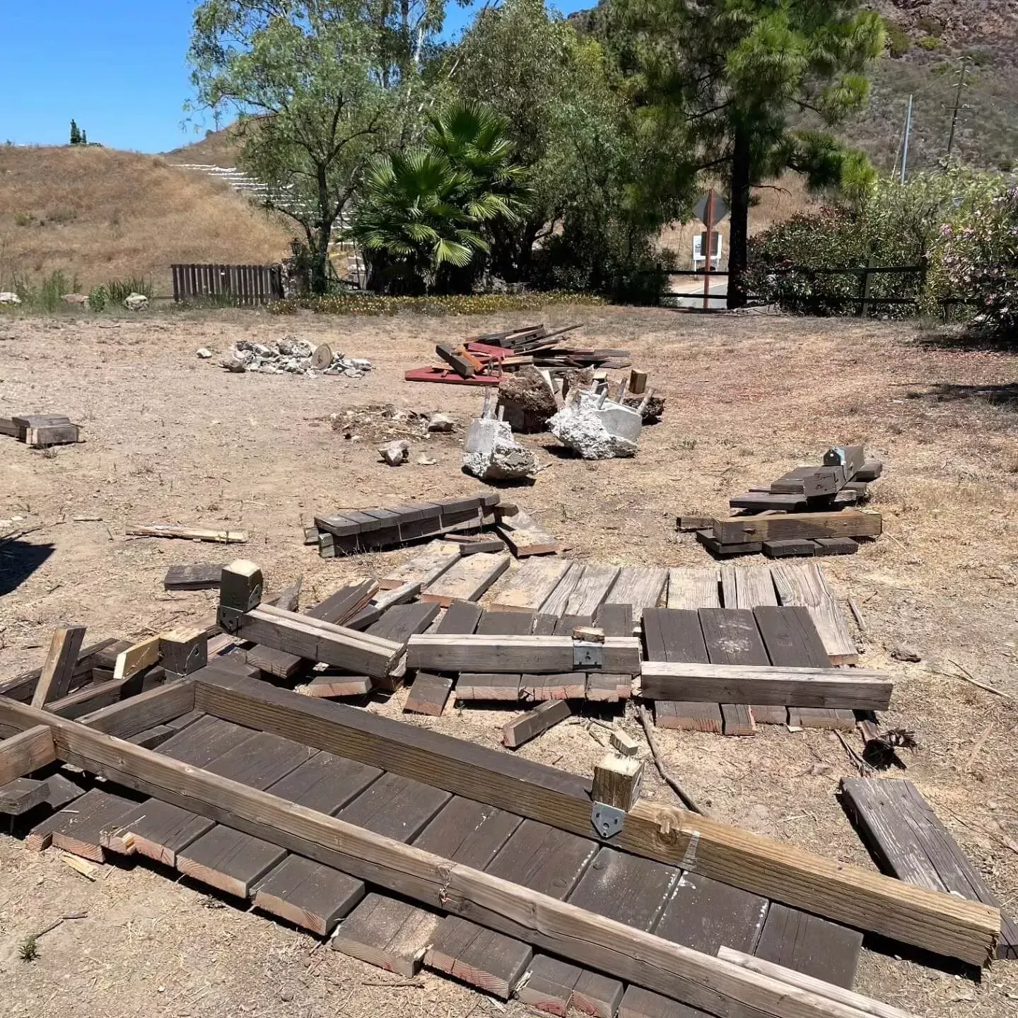 Construction materials scattered outdoors: wooden planks, beams, and bricks on a dirt ground, with trees and hills in the background.
