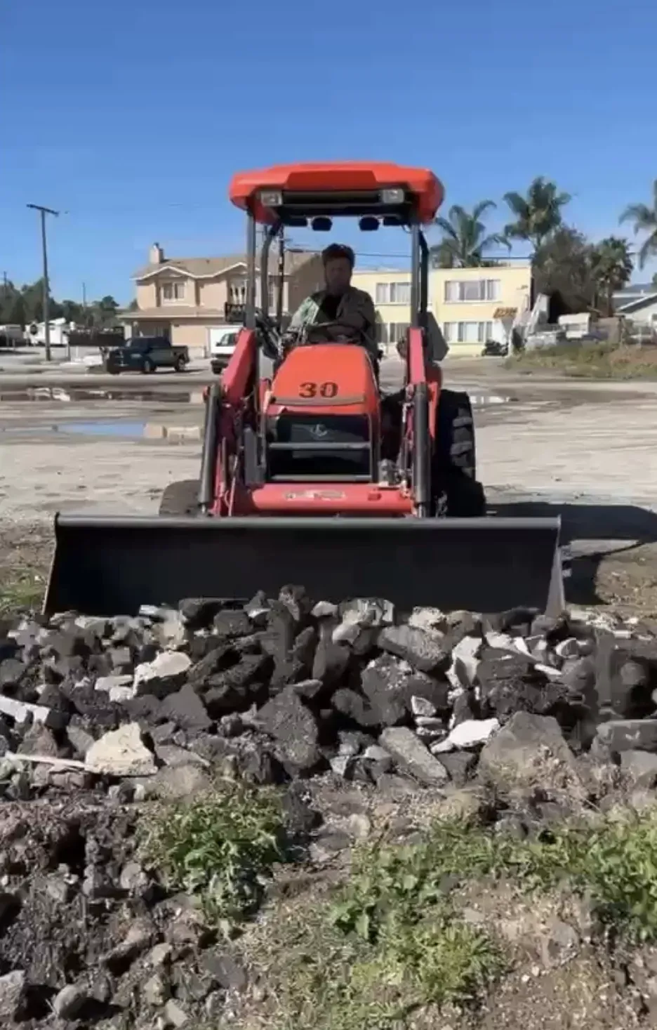 A person operates an orange skid-steer loader pushing a pile of dark rocks on a sunny, dirt-covered lot near houses.