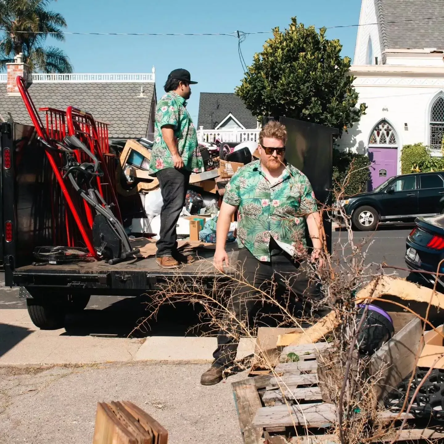 Two people in Hawaiian shirts loading a truck with debris, parked on a street. Buildings and a sunny sky in the background.