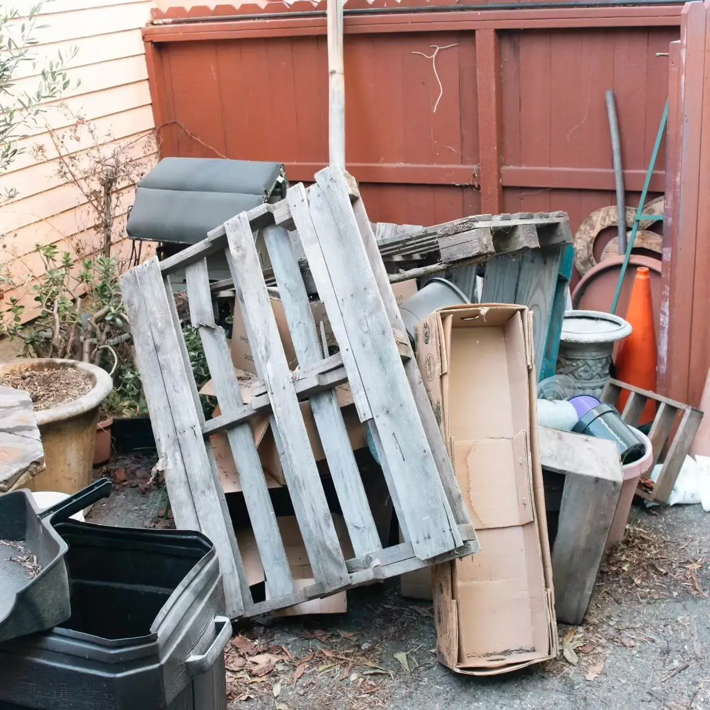 Pile of trash: wooden pallets, cardboard boxes, various debris next to a brown wooden fence.