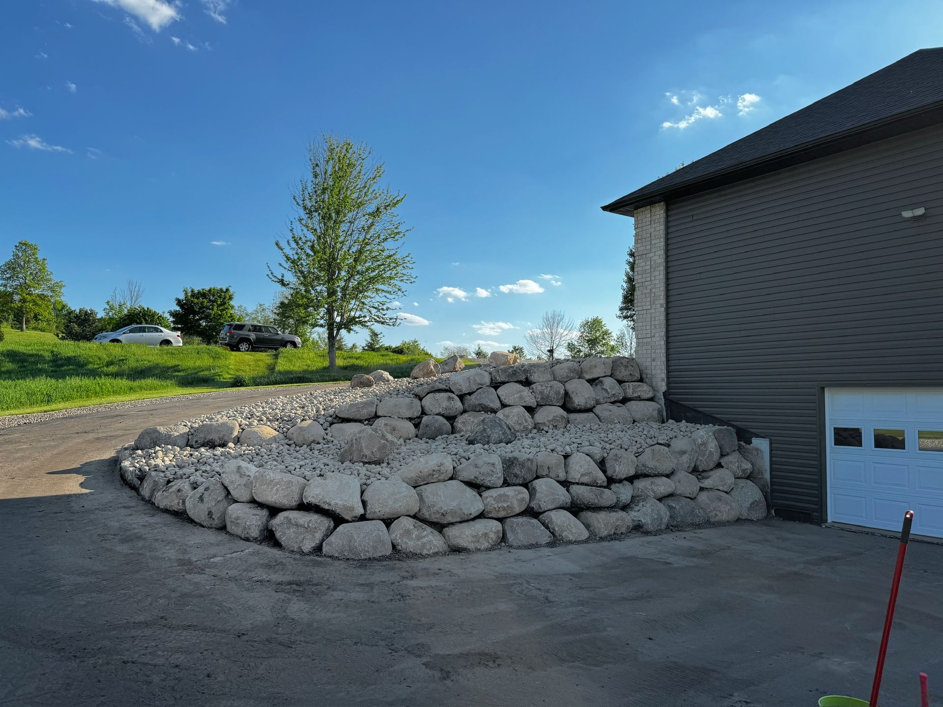 A rock retaining wall beside a building with a garage door, a gravel driveway, and a grassy hill under a blue sky.