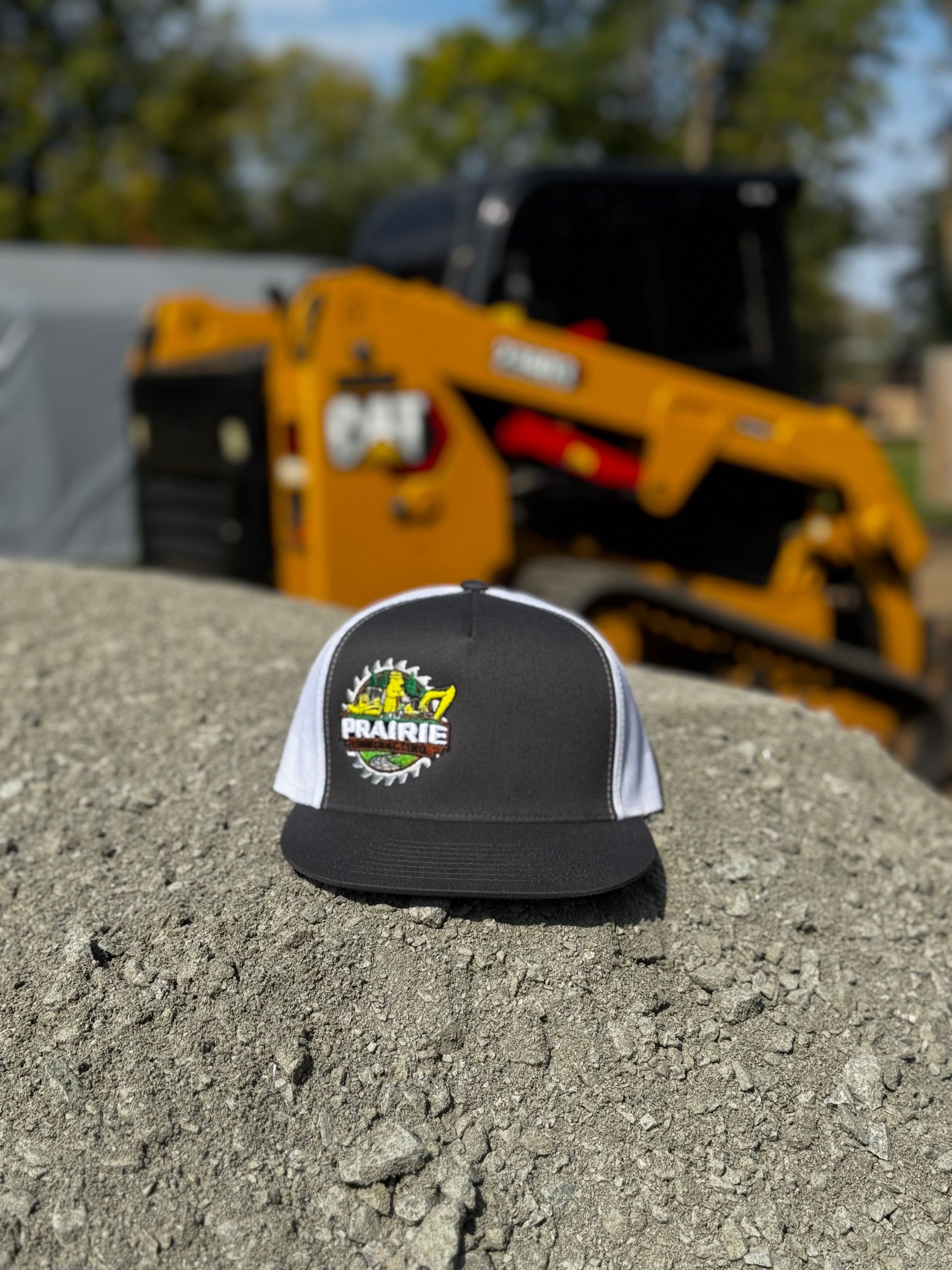 Black and white trucker hat with logo, on a pile of gravel, with a yellow skid steer loader in background.
