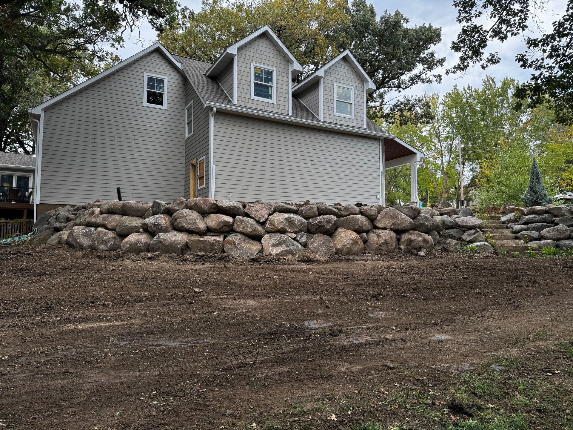 A light gray house with two dormers sits behind a large rock retaining wall on a leveled dirt lot.