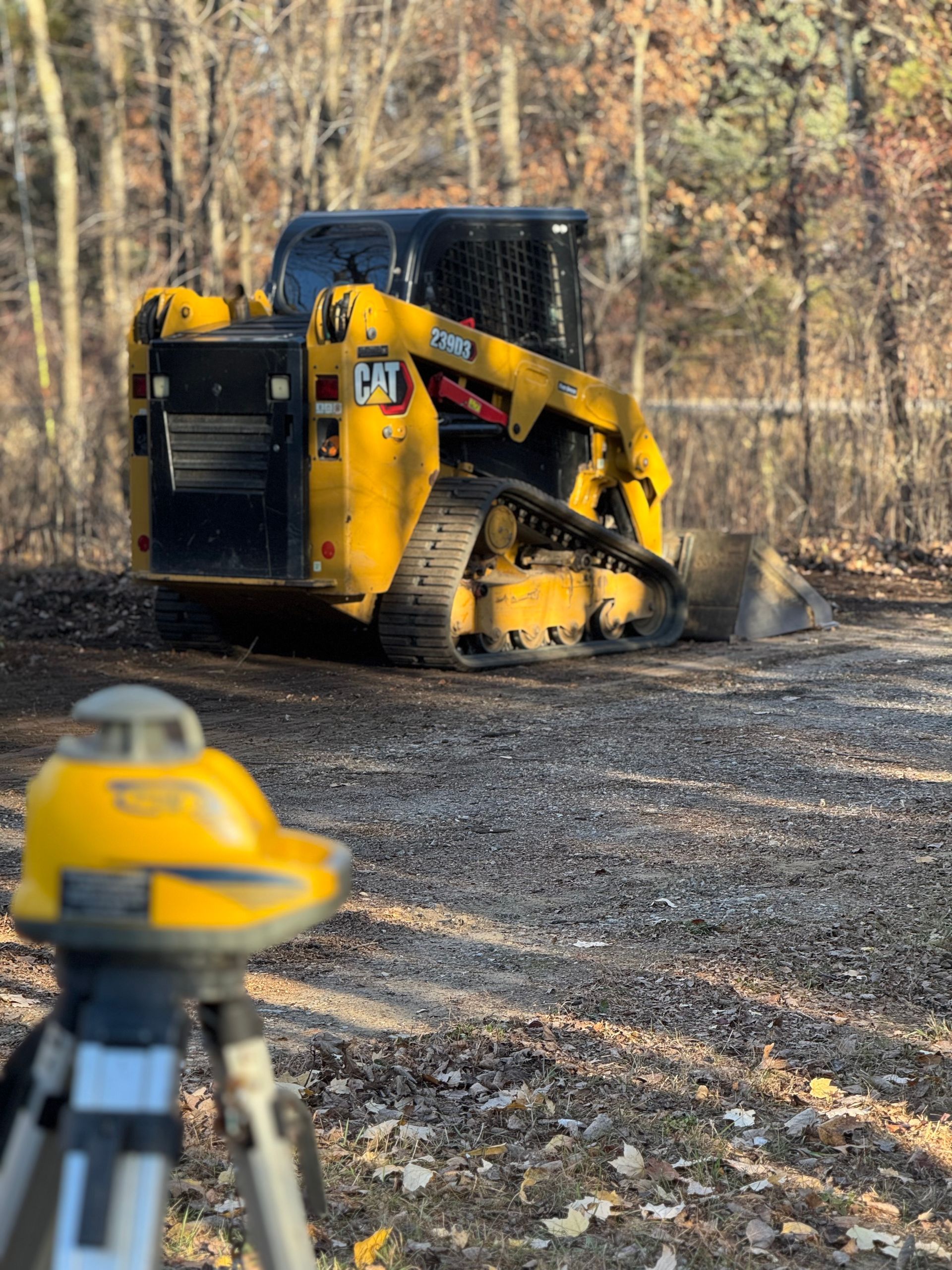 Yellow and black skid steer on gravel, with a surveying level in the foreground.