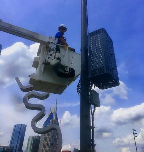 Worker in a lift bucket repairs equipment on a tall pole, city buildings in the background, sunny day.