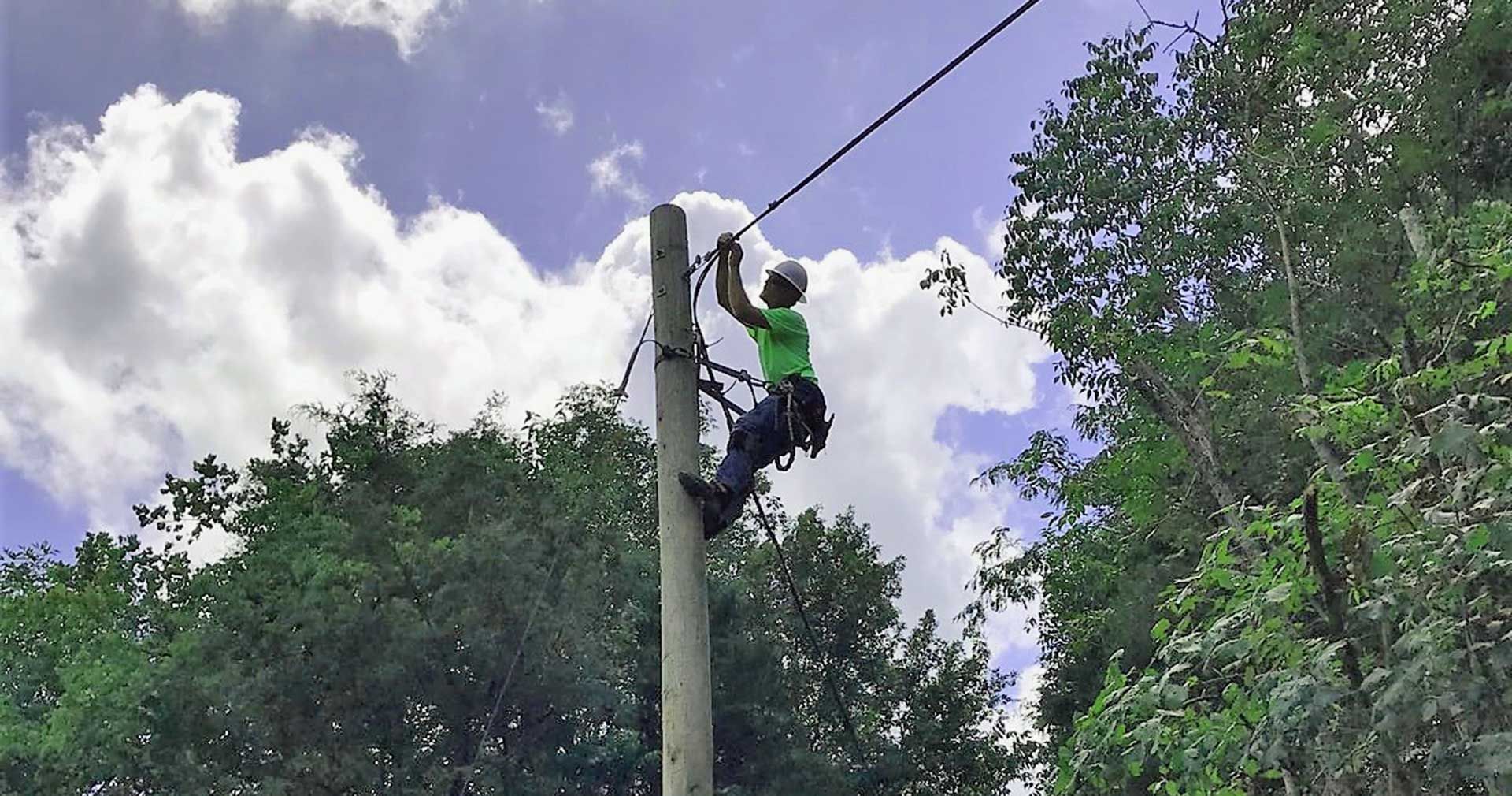 Lineman working on a power pole, wearing a green shirt and safety gear against a cloudy sky and trees.