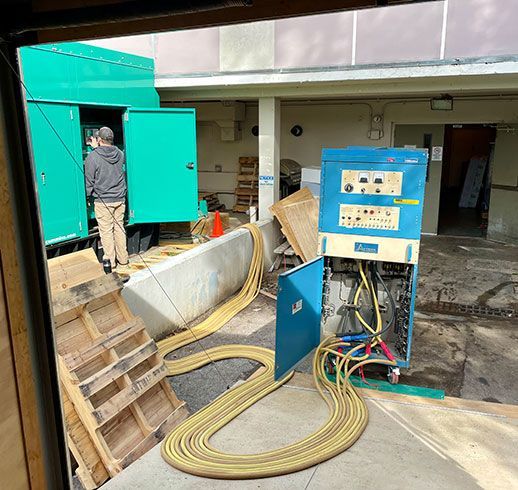 A person by a green generator truck, with yellow cables running to a blue power panel.