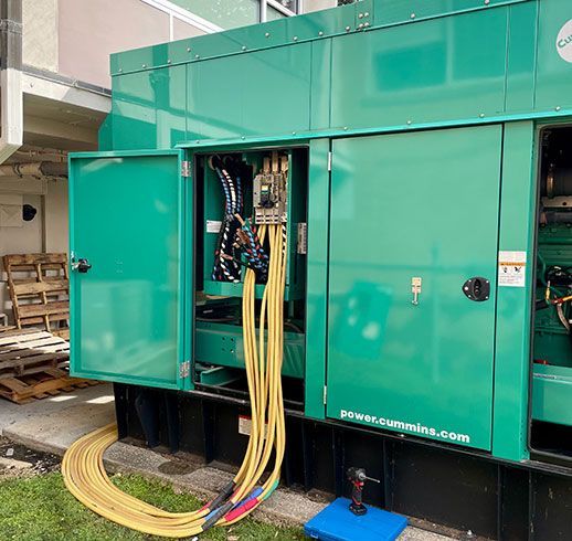 Green generator with open door and yellow cables, next to a building.