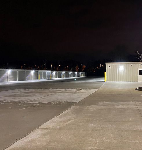 A nighttime view of a storage facility. Rows of storage units lit by bright lights. Empty parking area.