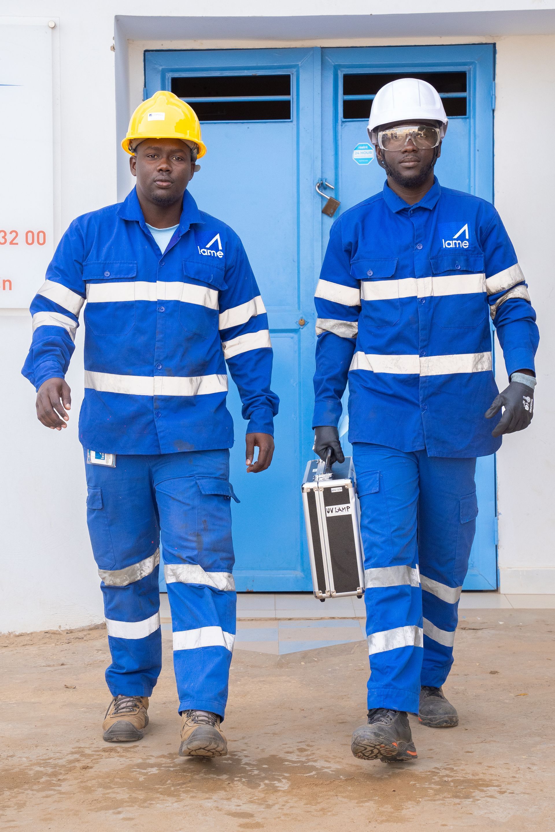 Deux hommes portant des salopettes bleues et des casques de chantier marchent devant une porte bleue.