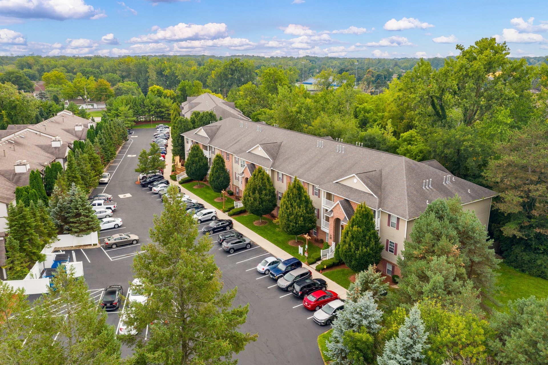 Aerial view of apartment complex with parked cars, surrounded by trees and green lawn.