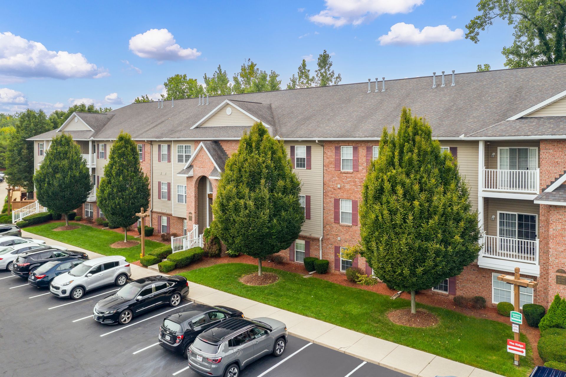Apartment building with cars parked in front. Red brick, beige siding, green trees, and blue sky.