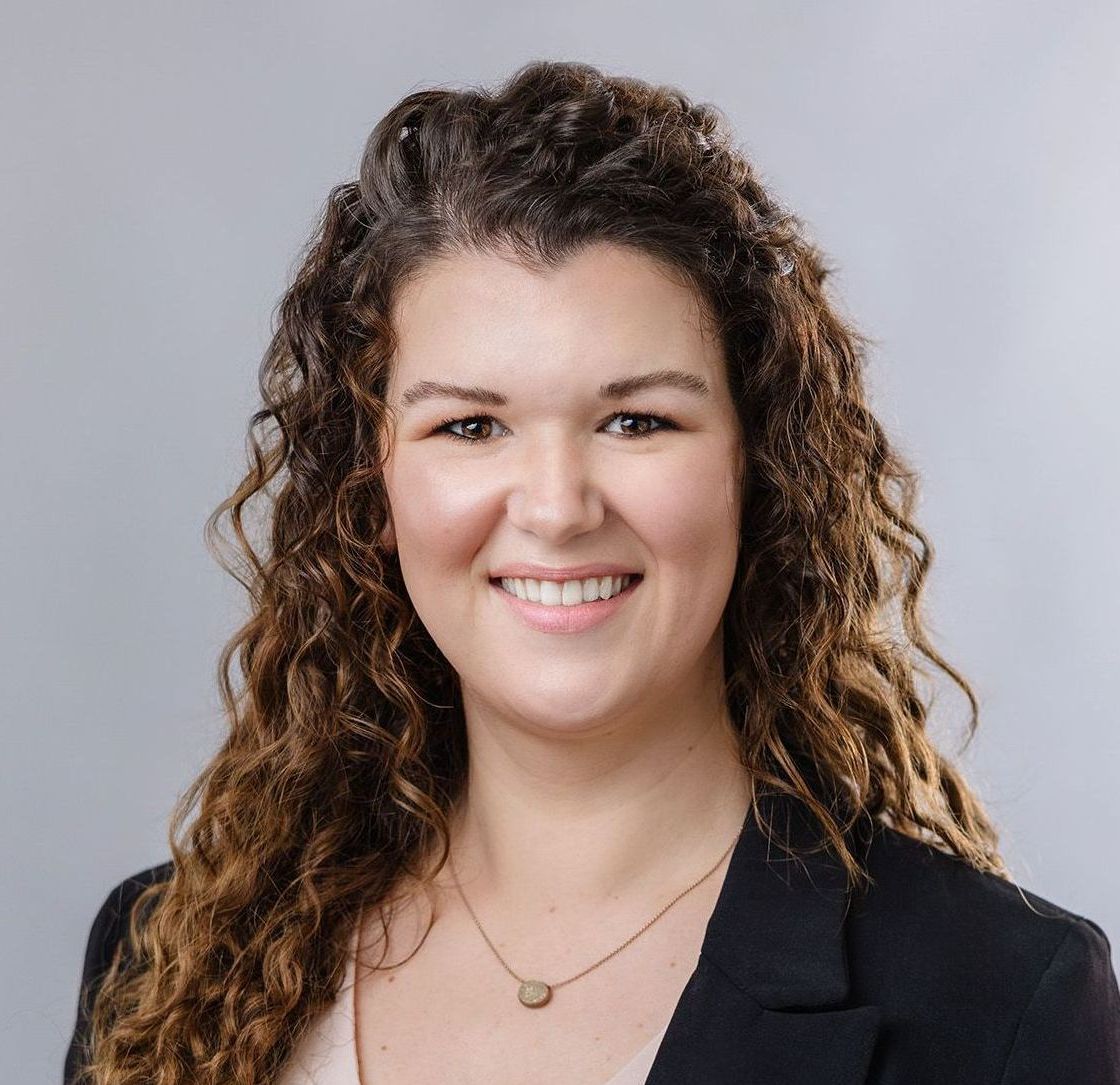 Kat has curly hair, is wearing a black jacket, and a necklace .