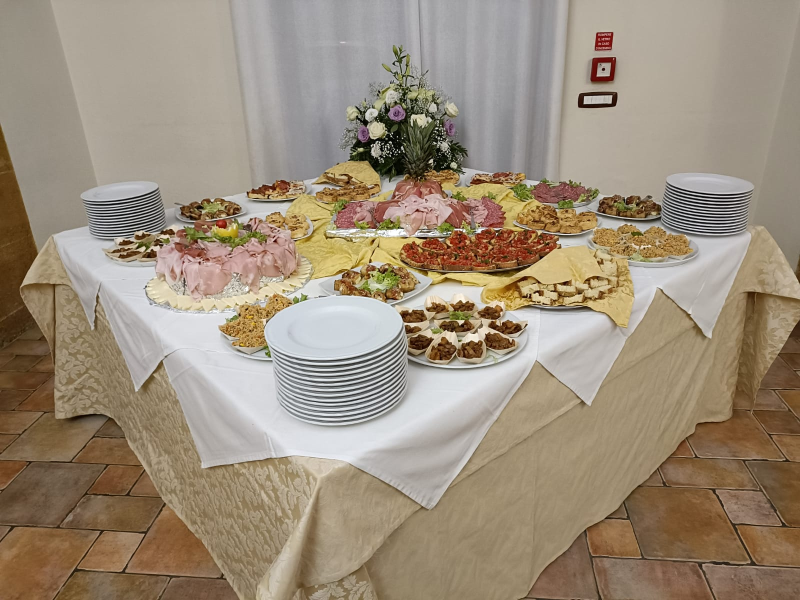 Tavolo da buffet con vari cibi: salumi, formaggi, antipasti. Tovaglia bianca, centrotavola floreale, piatti impilati.