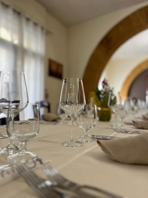 Lungo tavolo apparecchiato per un pasto con bicchieri da vino e posate, all'interno sotto un arco.