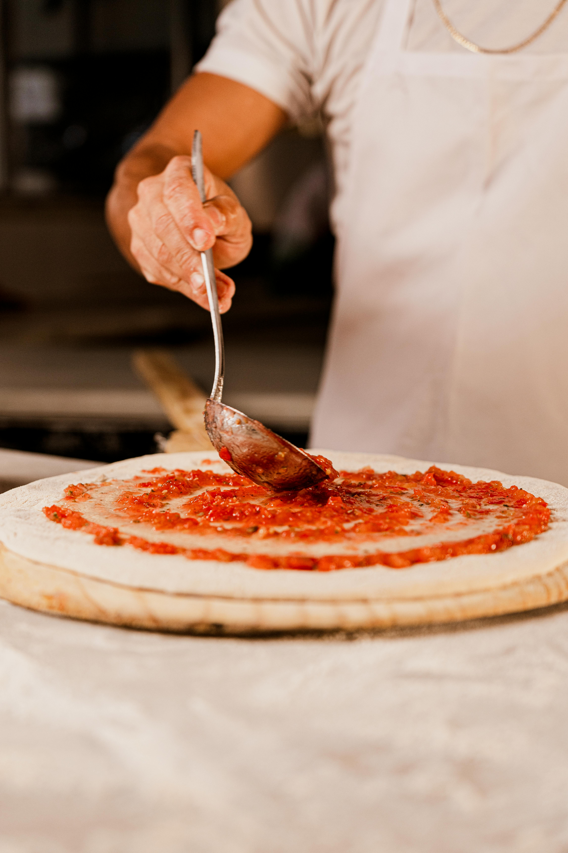 Chef che spalma la salsa di pomodoro sull'impasto della pizza con un mestolo.
