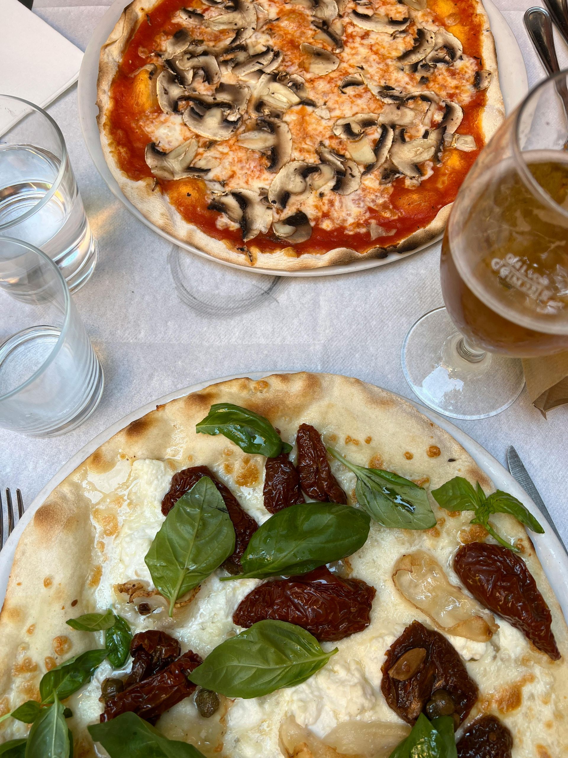 Due pizze, una con funghi, una con pomodori secchi e basilico, accompagnate da bicchieri d'acqua e birra.