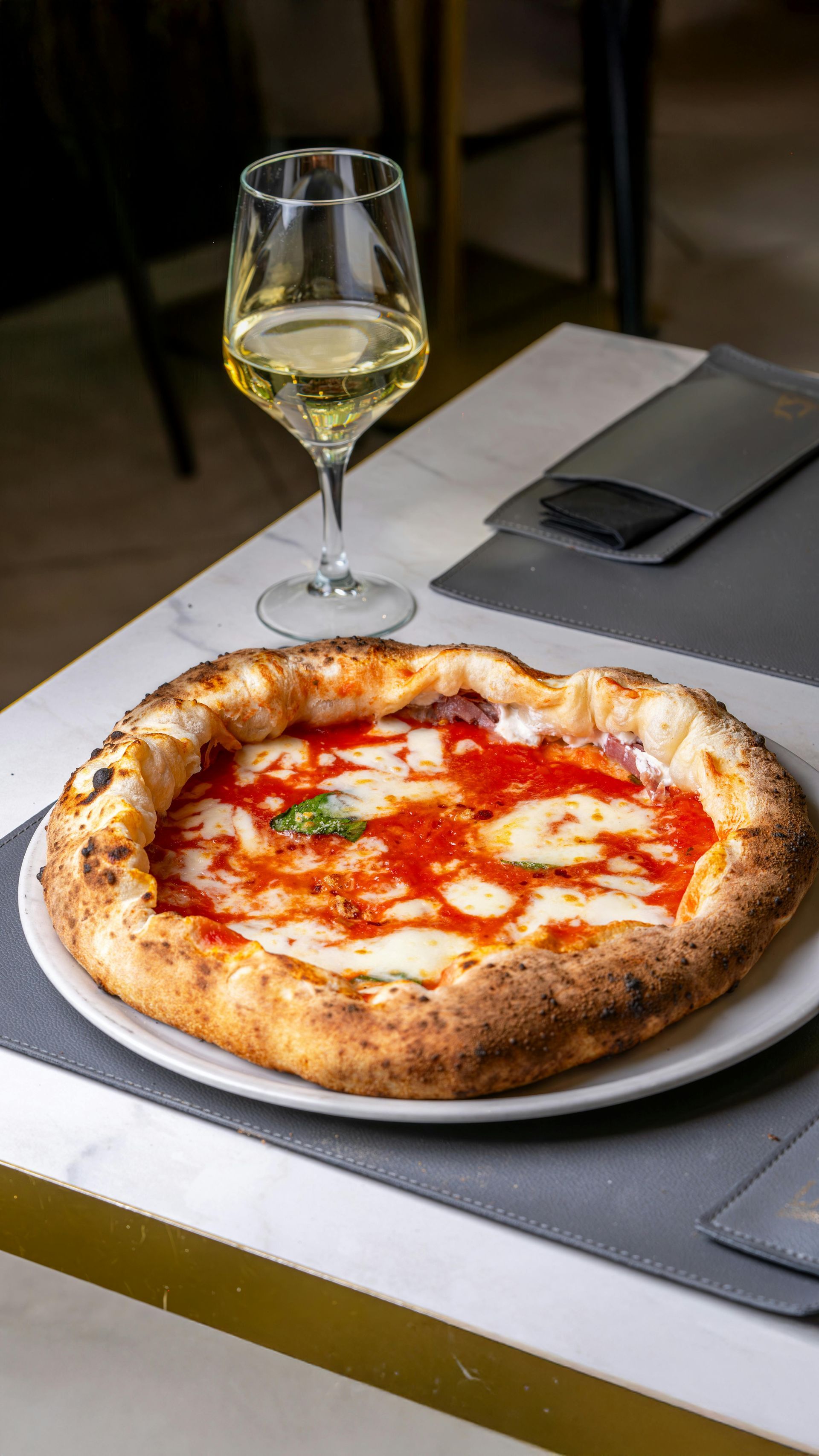 Pizza margherita su un piatto con un bicchiere di vino bianco sul tavolo.