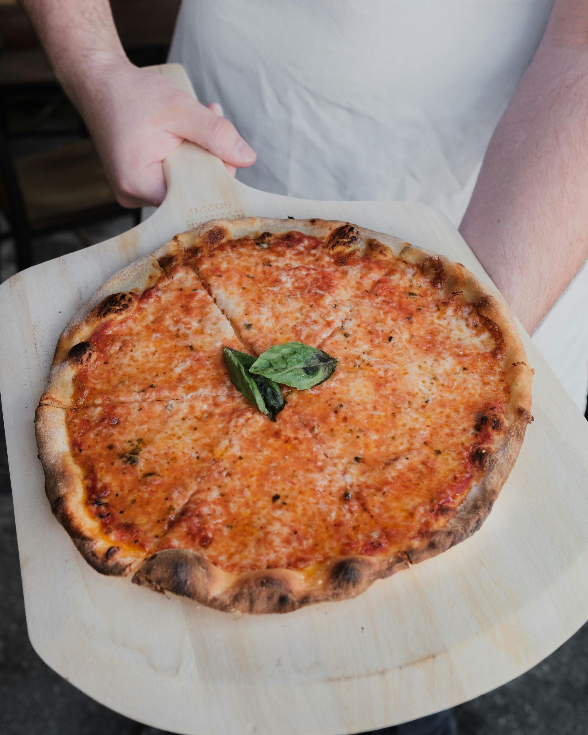 Pizza su una pala di legno, tenuta da una persona. La pizza è condita con salsa di pomodoro e formaggio, con una foglia di basilico sopra.