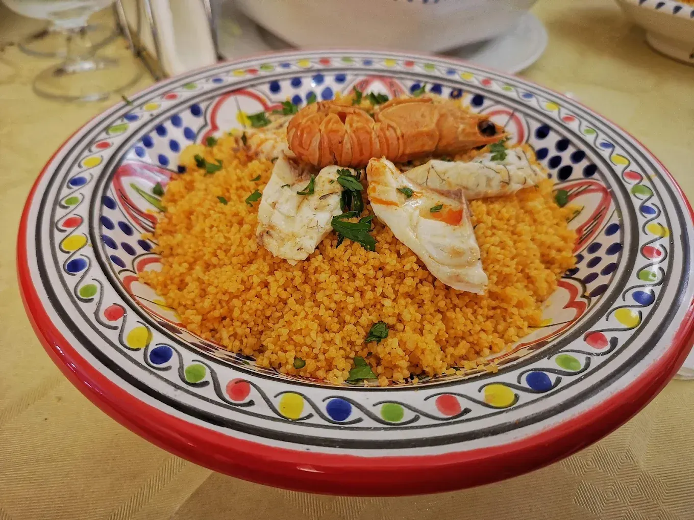 Couscous con gamberi e pesce, servito in una ciotola di ceramica decorata su un tavolo.