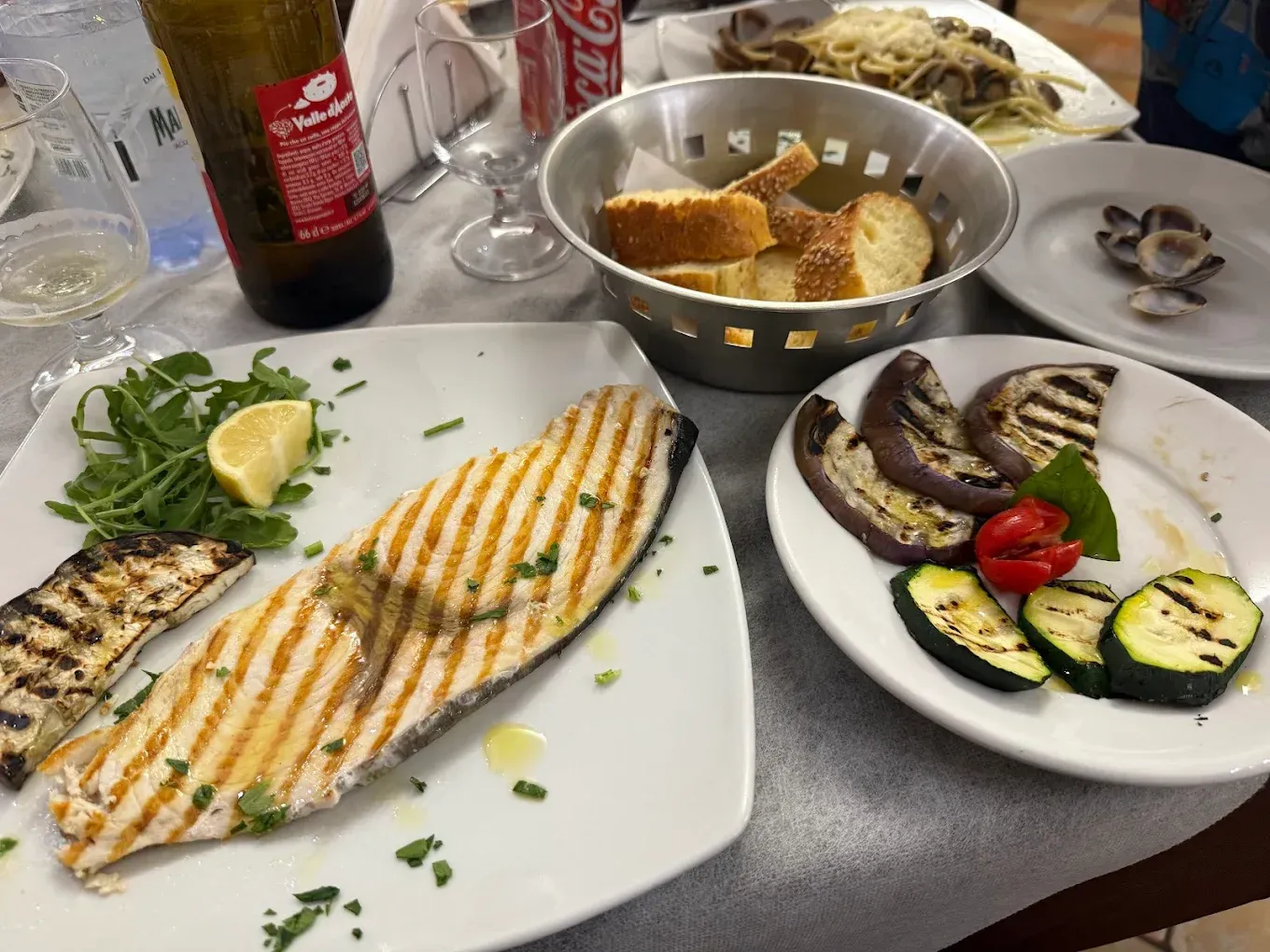 Pesce e verdure alla griglia con pane, spaghetti e vongole su un tavolo di un ristorante.