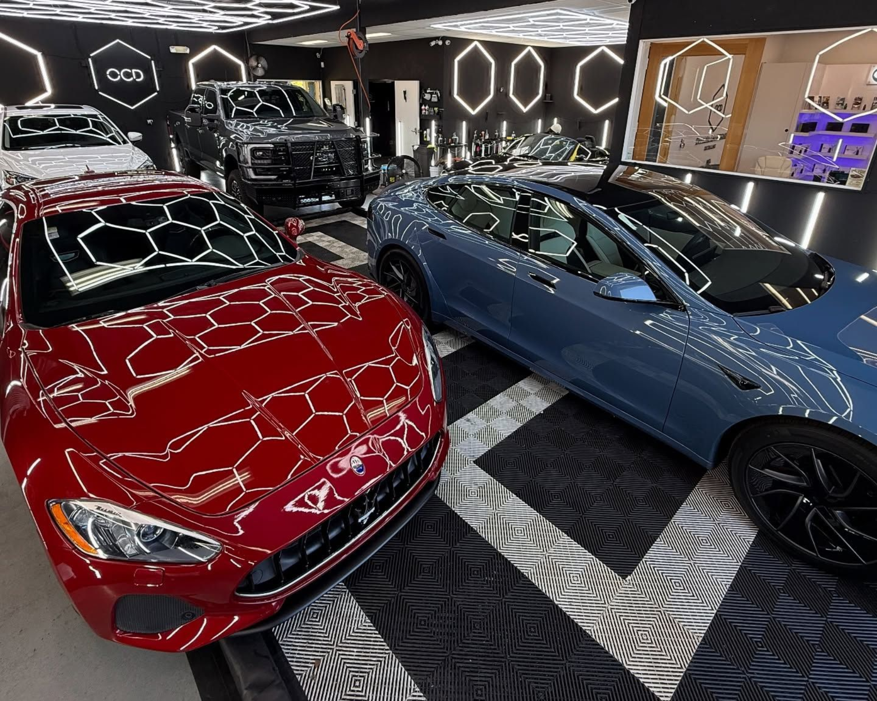 Red sports car and blue Tesla in a showroom, with other vehicles and hexagonal lighting.