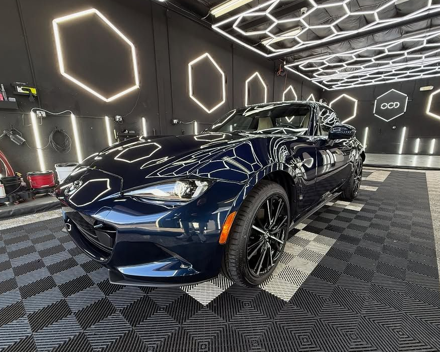 Dark blue Mazda MX-5 Miata in a car detailing bay with hexagonal lights and checkered flooring.