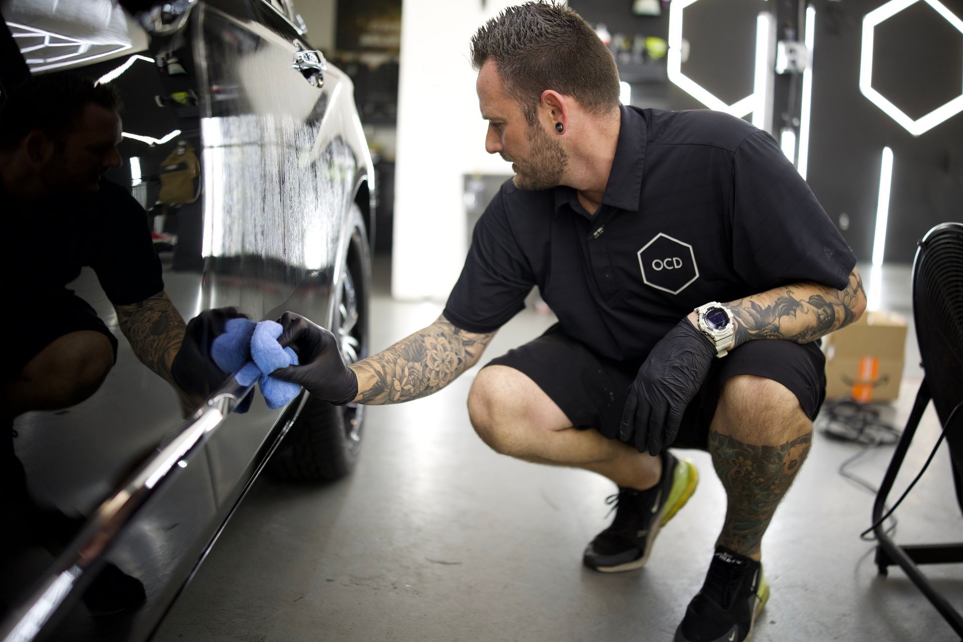 Man wiping the front grille of a black car with a cloth and sponge in a garage setting.