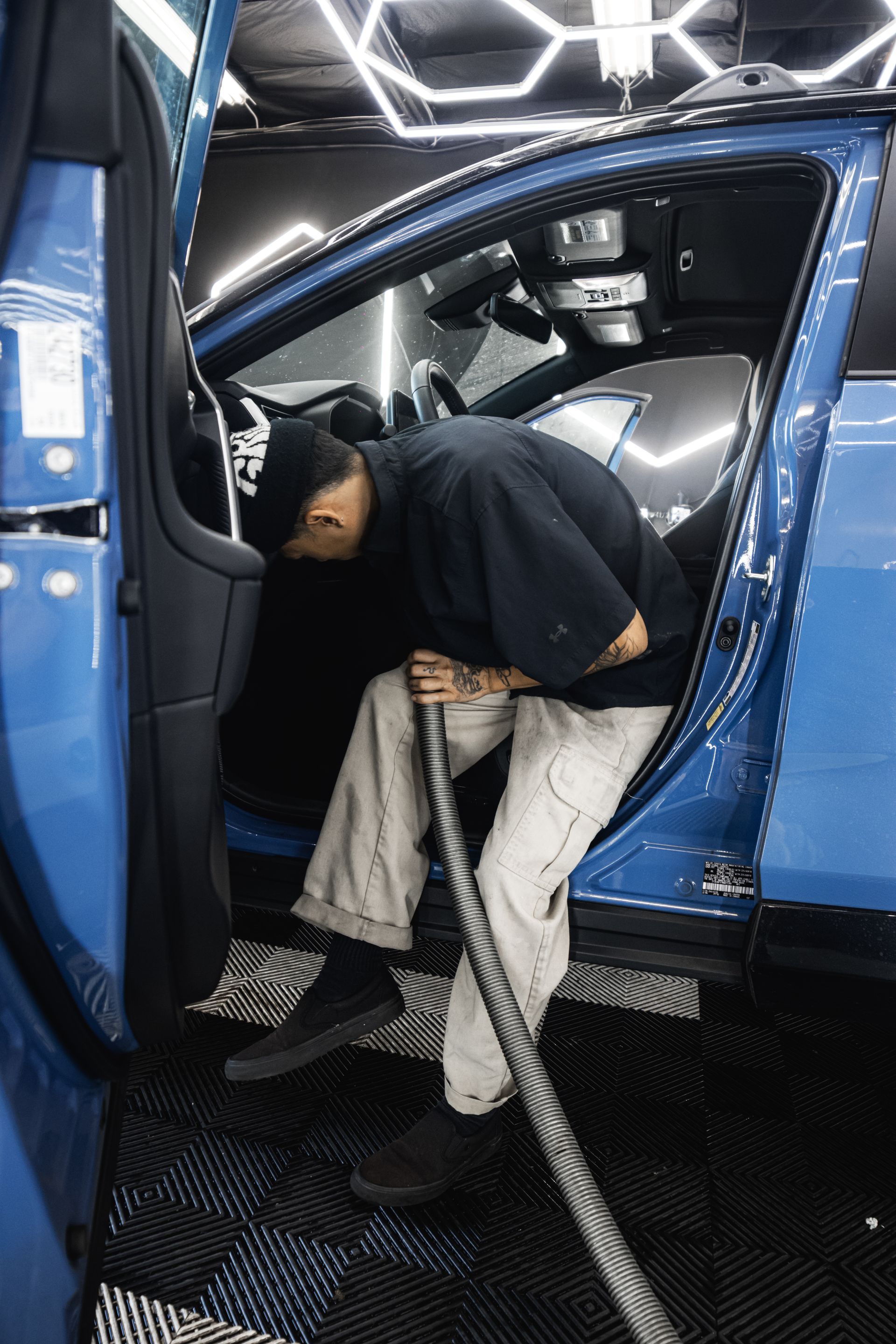 A person vacuums the interior of a blue car. They are inside the open door, using a long hose.