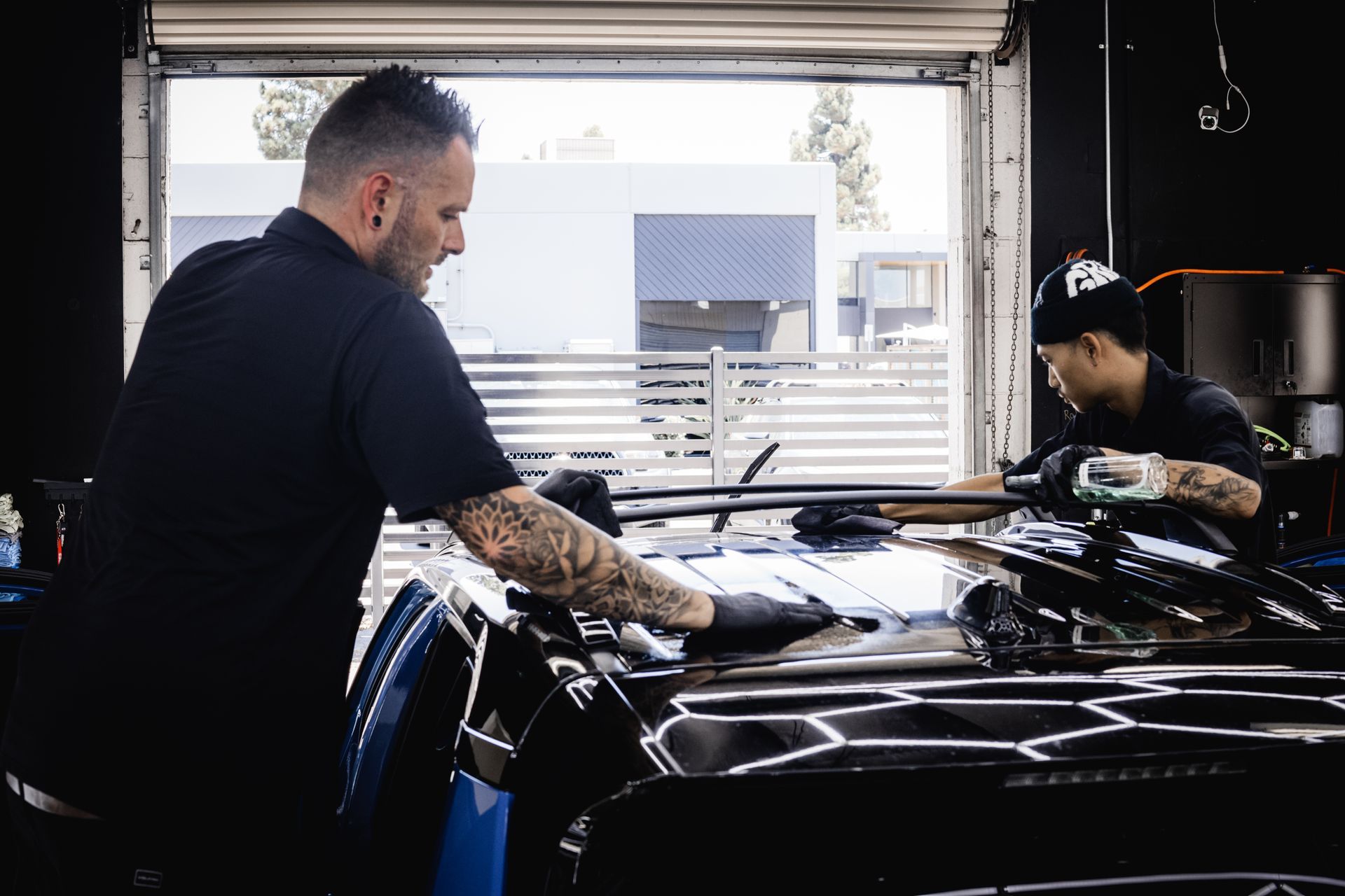 Two people detailing a black car in a garage with large windows. One is applying something; the other is using a machine.
