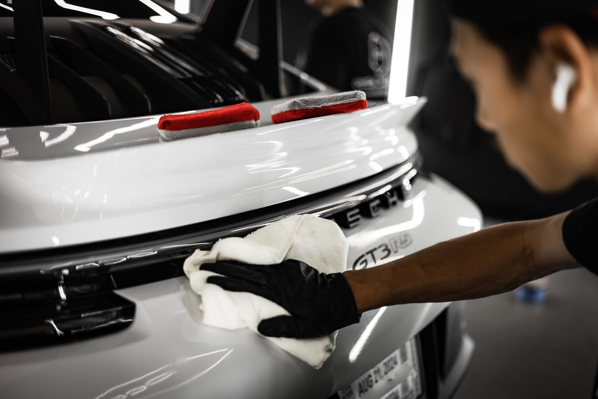 Person wearing black gloves wiping a white Porsche GT3 with a microfiber cloth in a brightly lit garage.