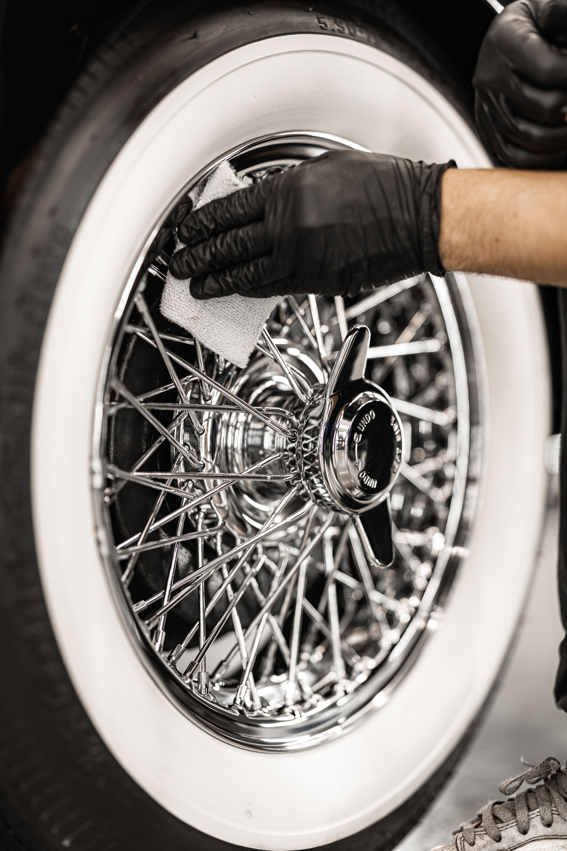 Person wearing black gloves cleaning a classic car's wire wheel with a white cloth.