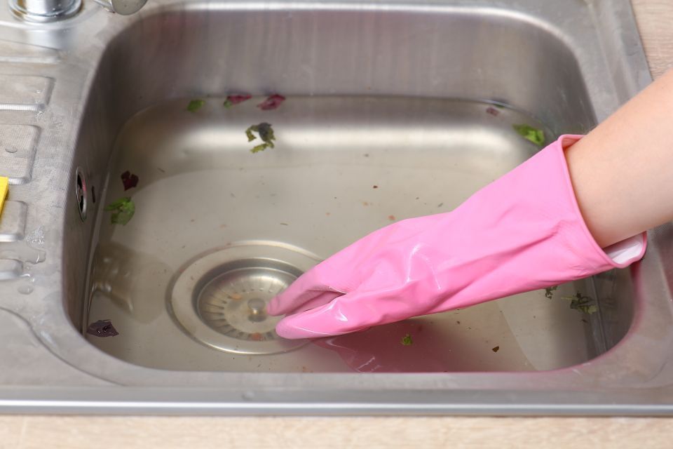 Pink-gloved hand reaches into a partially filled kitchen sink to clear debris near the drain.