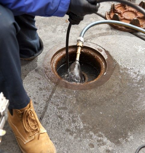 Person in work boots, blue jacket, and black gloves cleaning a sewer drain with a hose - commercial plumber barossa valley