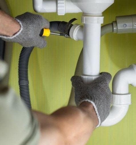 Hands in work gloves adjusting white PVC plumbing under a sink. Yellow screwdriver visible - residential plumber barossa valley