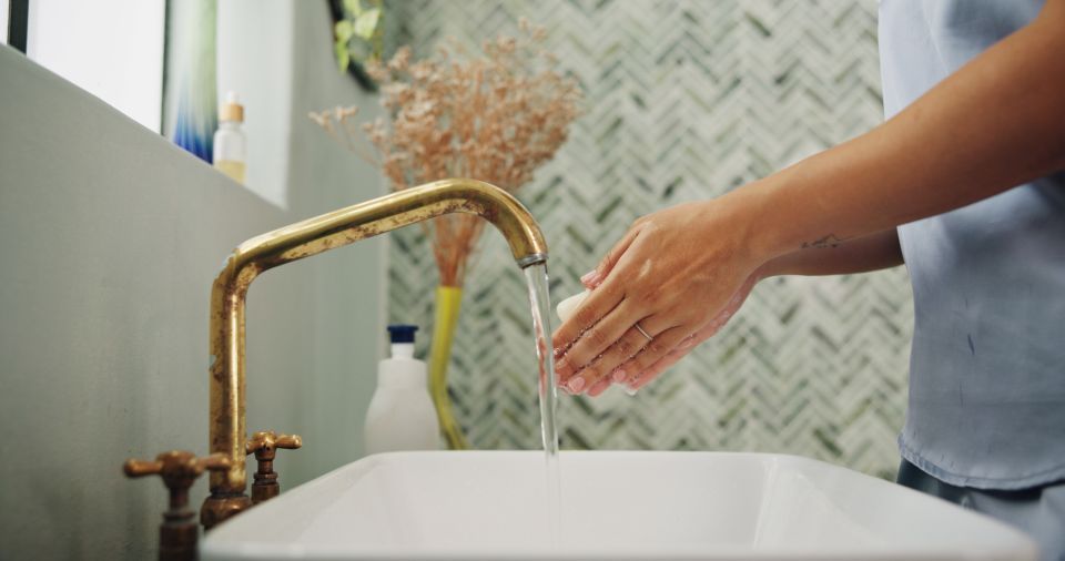 Person washing hands in a white sink under a gold faucet. Beige and green tiled wall in background.