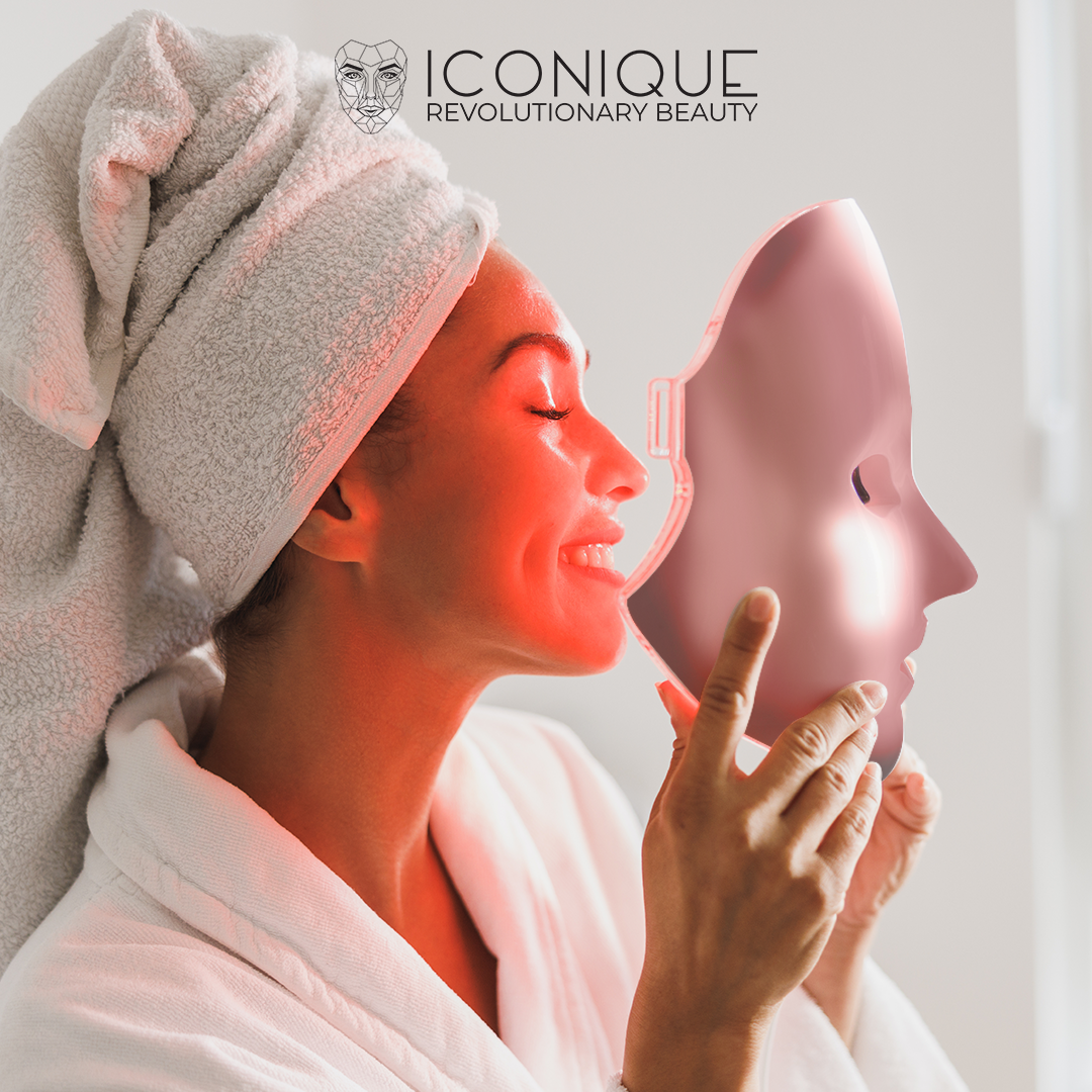 Frau im weißen Bademantel und Handtuch lächelt und hält eine rosa LED-Gesichtsmaske; rotes Licht leuchtet auf ihr Gesicht.