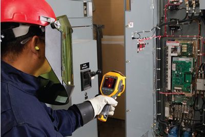 Electrician using a thermal imager on an electrical panel, wearing safety gear in an industrial setting.