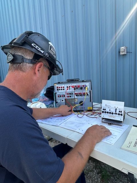 Person in a hard hat inspecting electrical equipment on a table with diagrams. Blue building background.