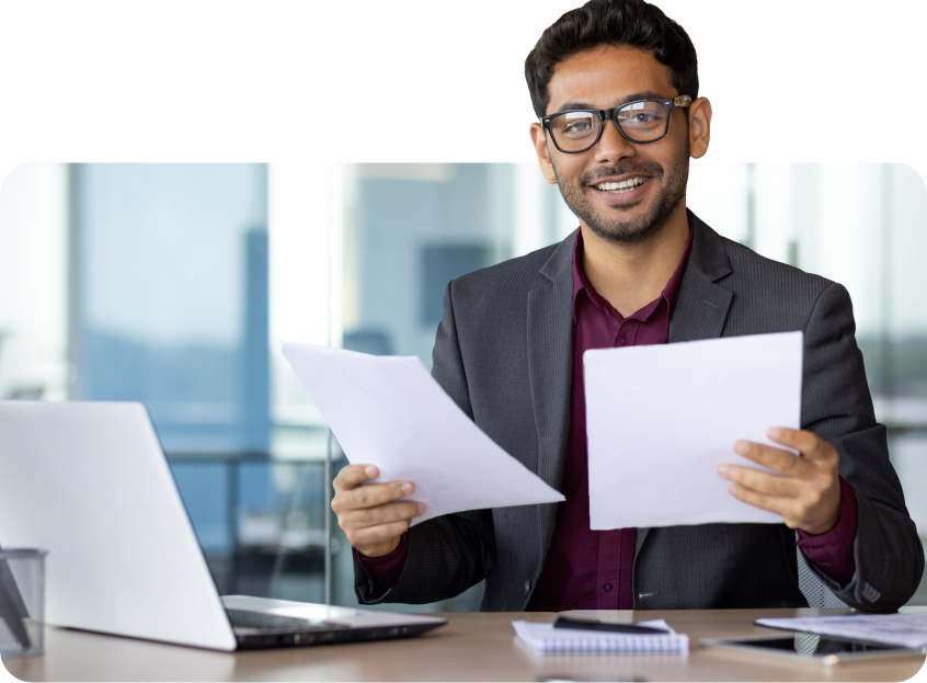 Um homem está sentado em uma mesa segurando papéis e um laptop.
