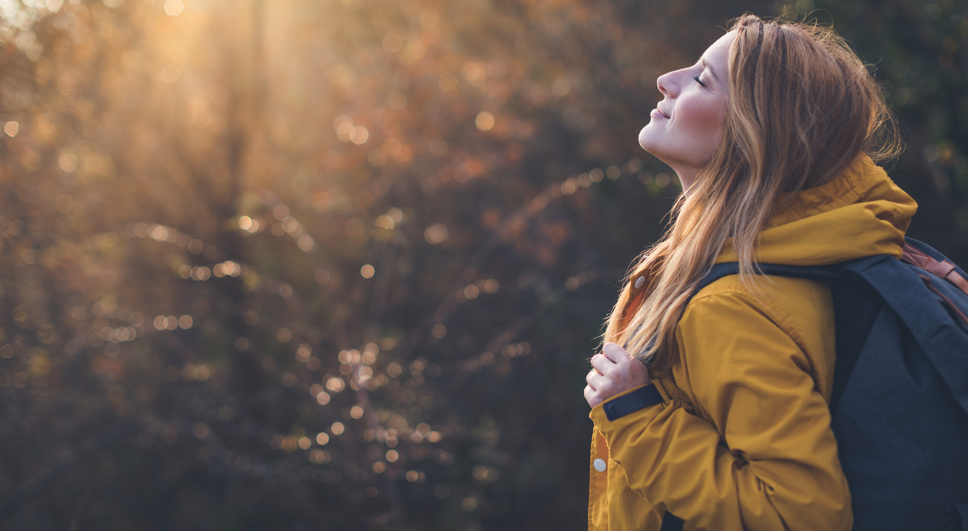 Woman with blonde hair, in a yellow jacket, looks up at the sunlight while hiking in a forest with a backpack.