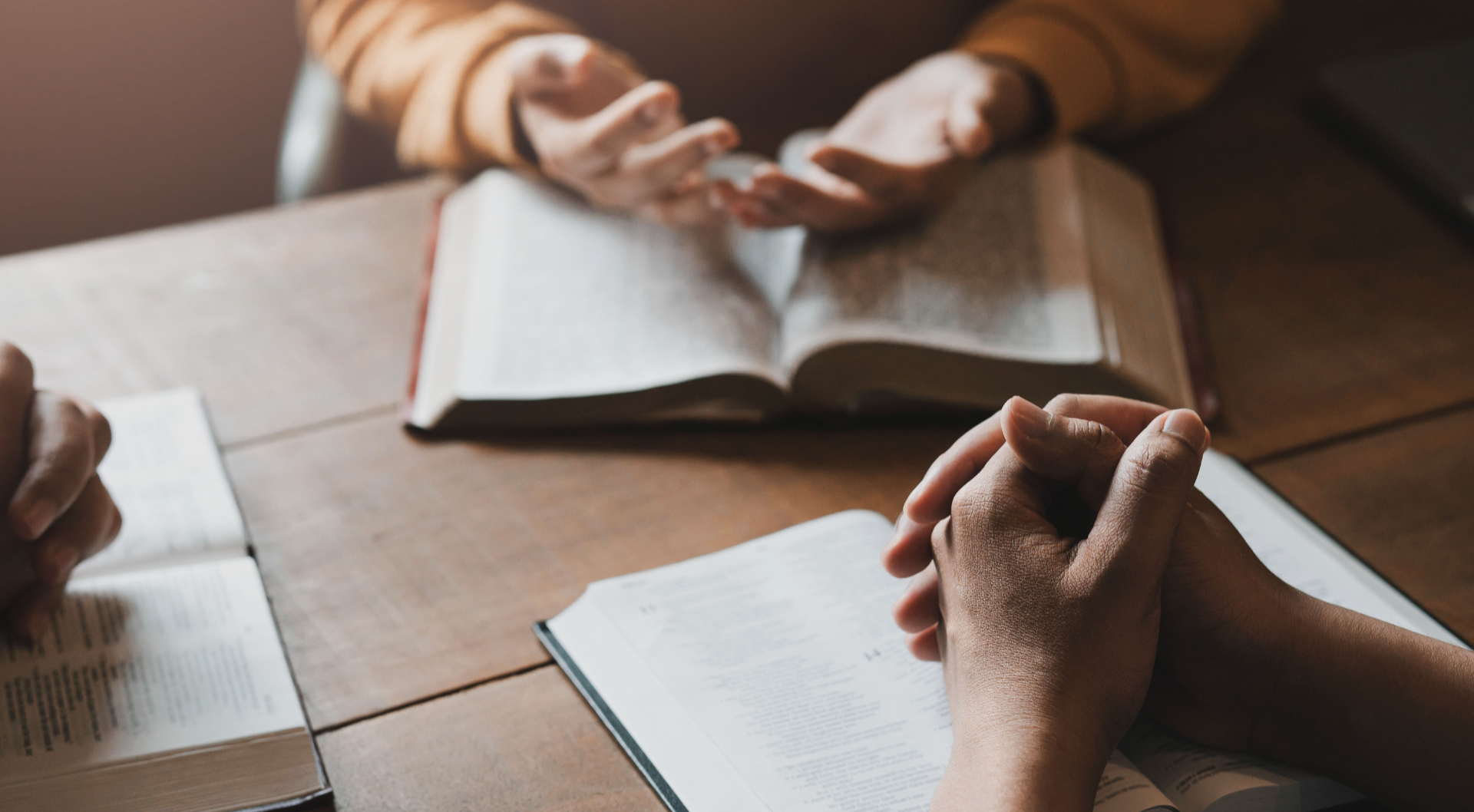 Hands clasped in prayer around open books on a wooden table, likely a Bible study or prayer meeting. Warm lighting.